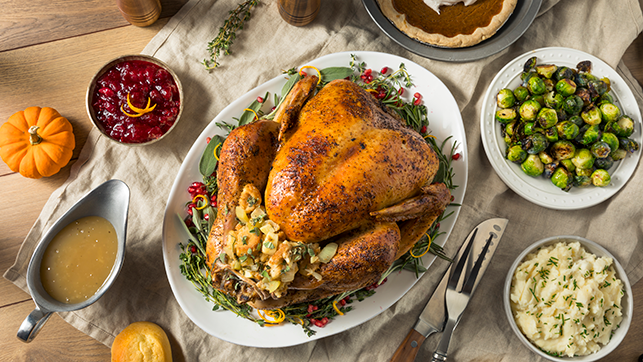 Roasted turkey on a white platter garnished with herbs and stuffing, surrounded by traditional Thanksgiving sides including mashed potatoes, Brussels sprouts, cranberry sauce, gravy, a dinner roll, and a pumpkin pie.