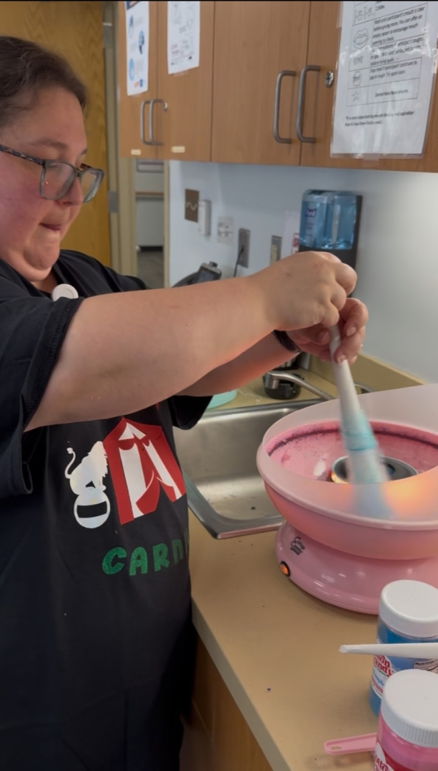 A person makes cotton candy using a pink machine on a countertop, spinning a paper cone to collect the candy. They wear a black apron with a circus tent graphic. Wooden cabinets and containers of floss sugar are visible in the background.